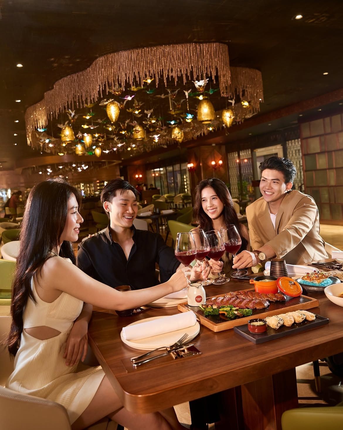 Một nhóm bạn nâng ly vang đỏ dưới đèn chùm, bàn gỗ bày đầy món nướng